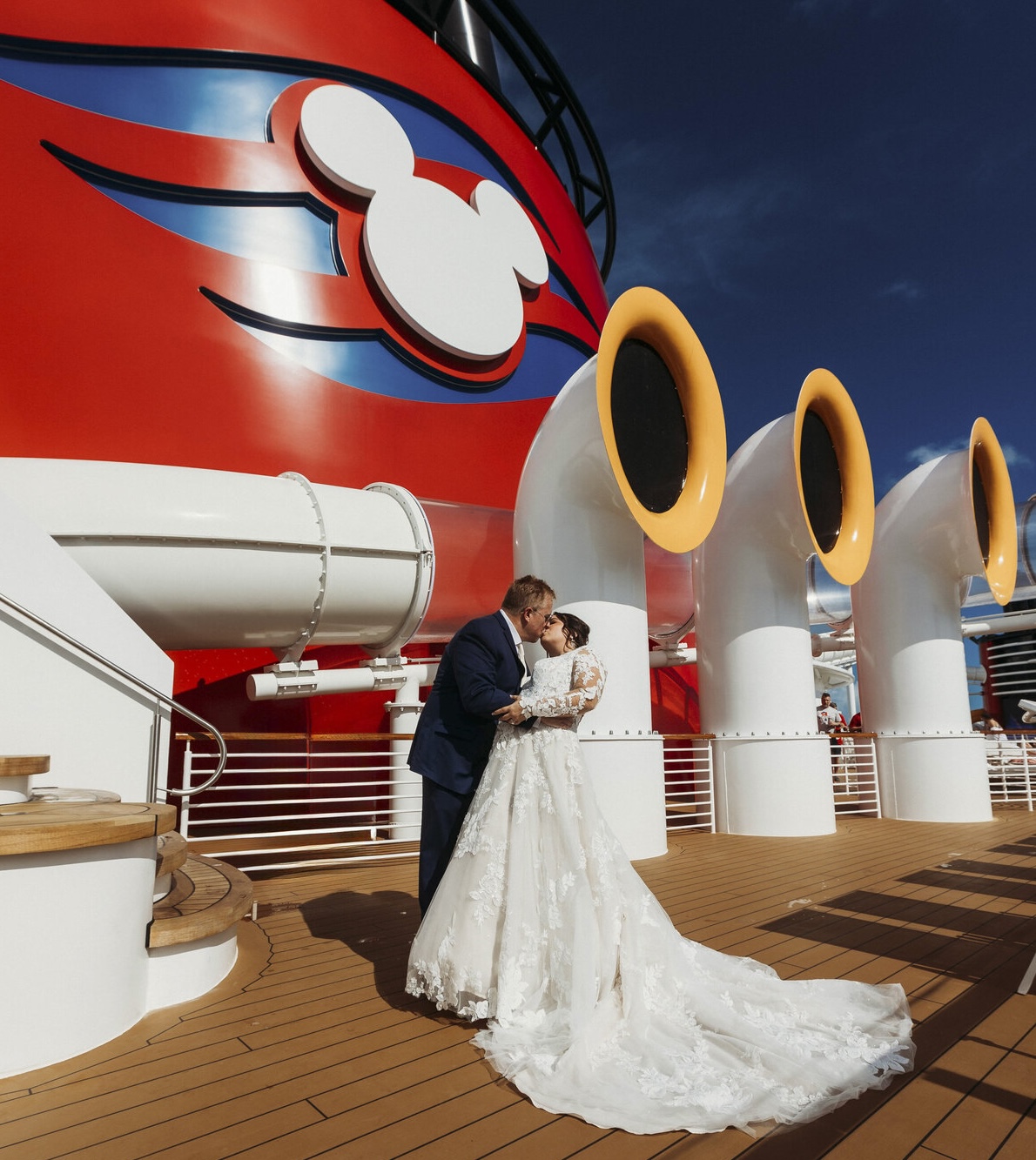 Bride and groom kissing on the deck of the Disney Wish in front of the red funnel, with the bride in a lace gown with a long train